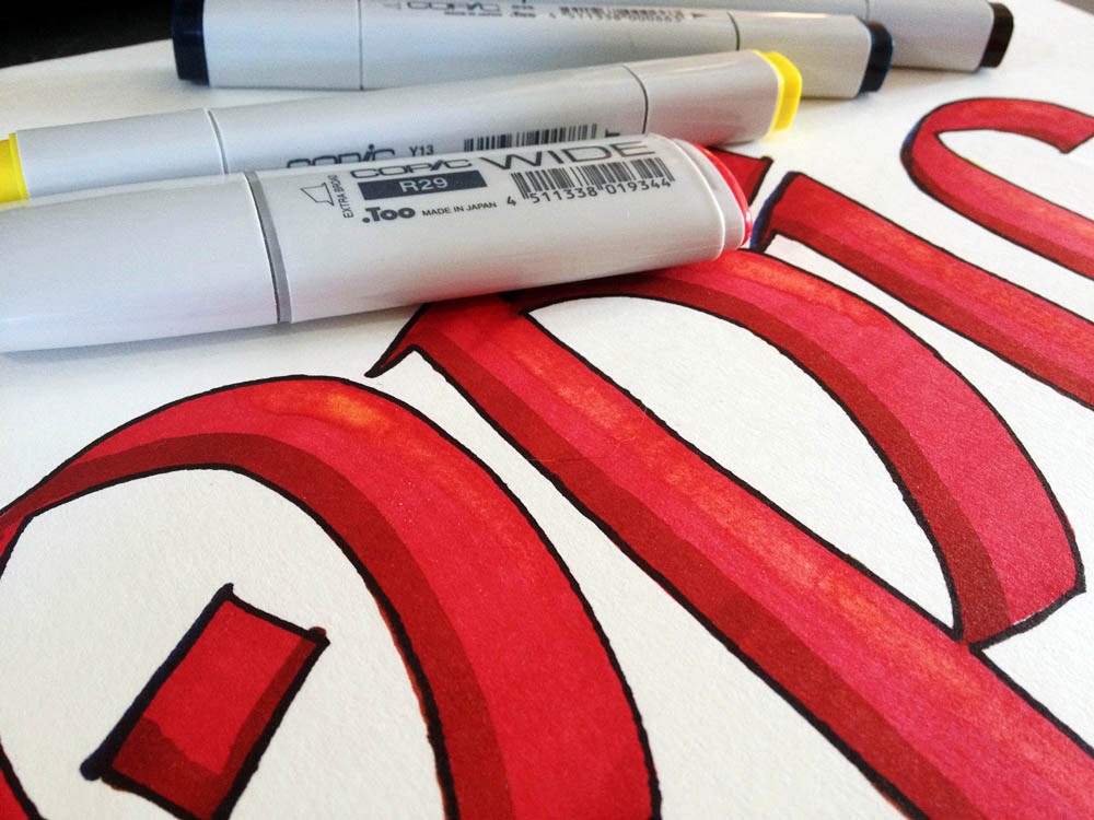 Copic Wide Marker liegen auf einem handgezeichneten roten Copic-Schriftzug auf hellem Papier.