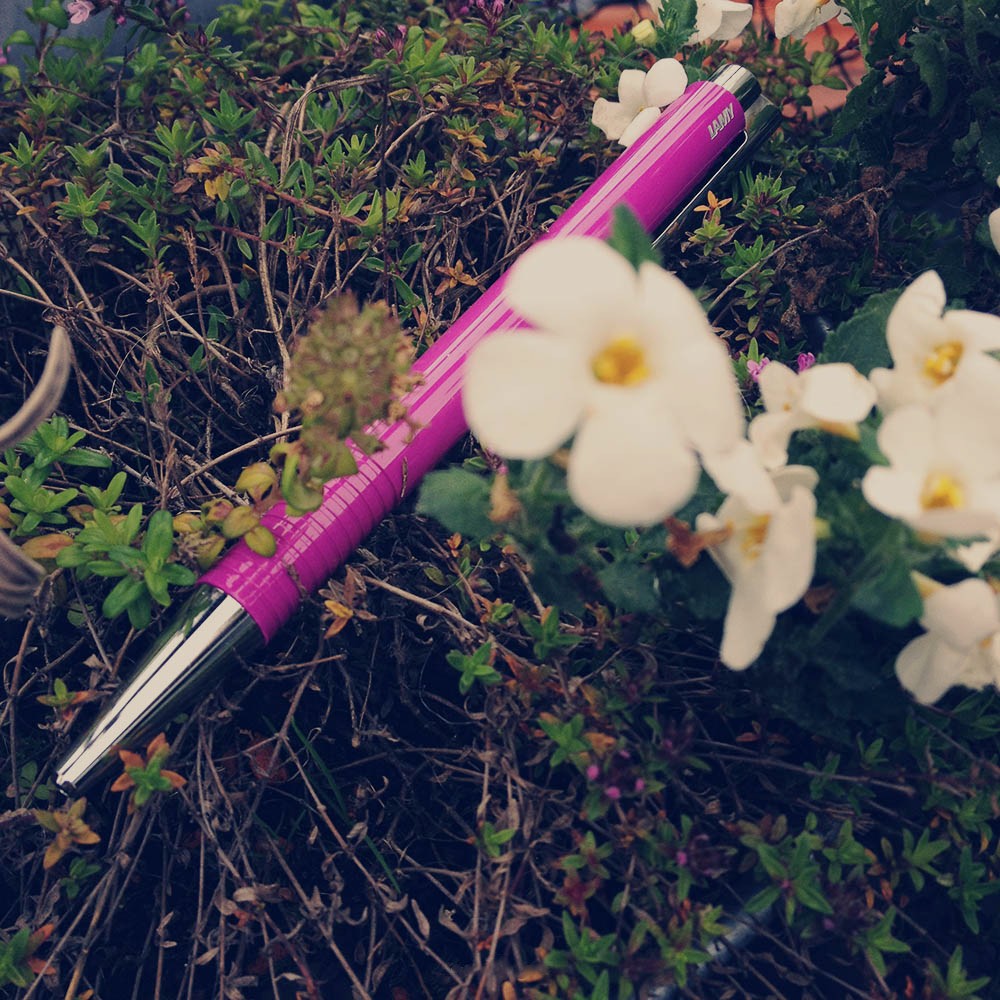 Violetter LAMY logo M+ Kugelschreiber zwischen weißen Blüten und grünen Pflanzen.