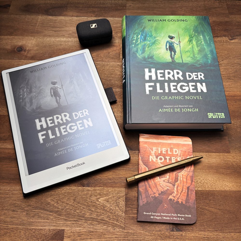 PocketBook InkPad One neben der Graphic Novel Herr der Fliegen, einem Notizheft und einem Messingstift auf einem Holztisch.