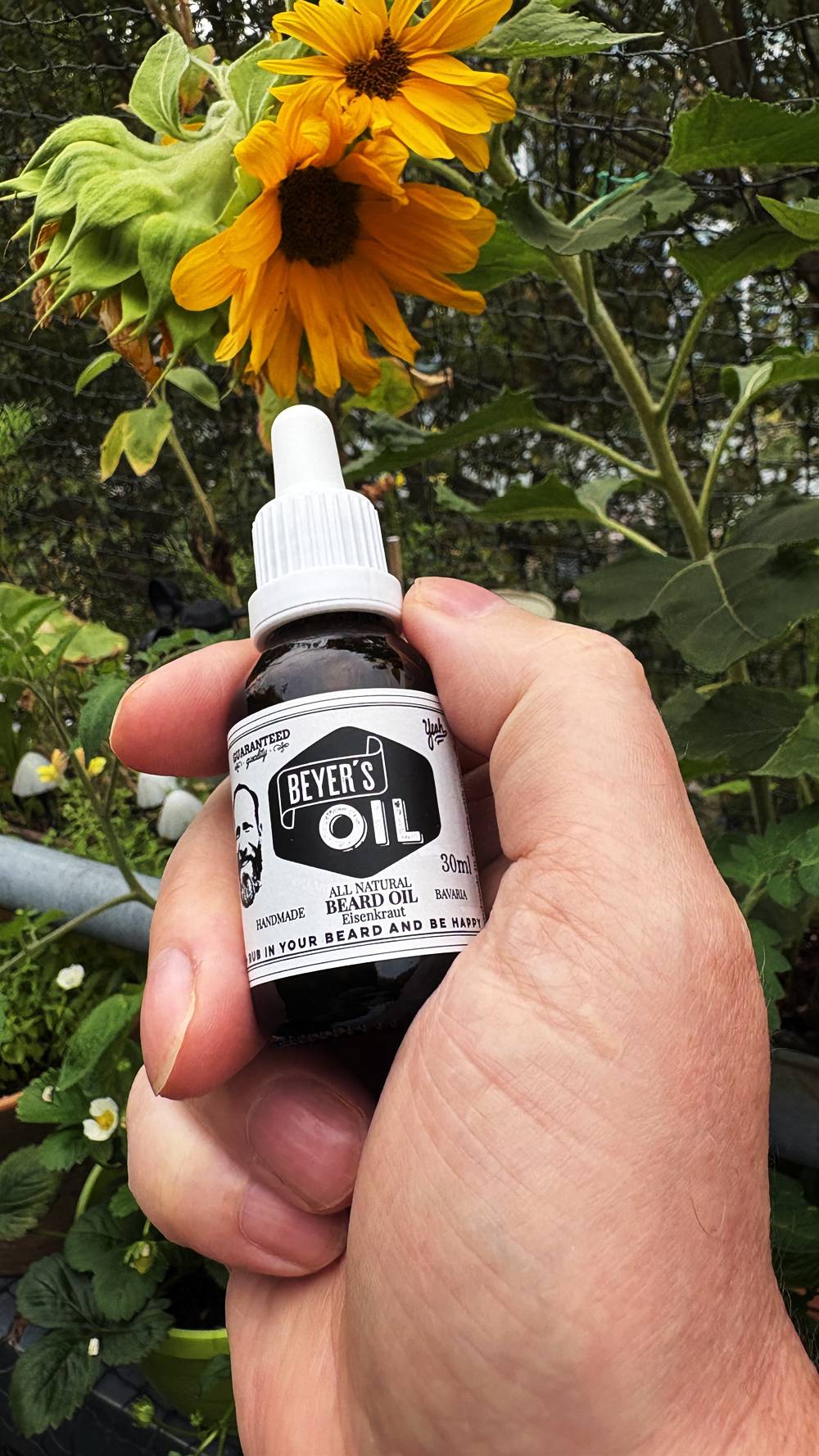 Flasche Beyer’s Oil Bartöl Eisenkraut in der Hand vor Sonnenblumen